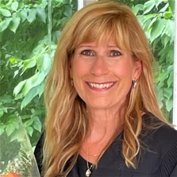 Head and shoulders bio pic of white woman, fair haired, smiling, wearing dark shirt, necklace, in fore and outside forest greens in the background.